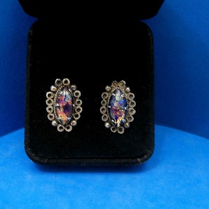 May include: A pair of silver earrings with iridescent glass stones set in a circular design. The earrings are displayed in a black velvet box.