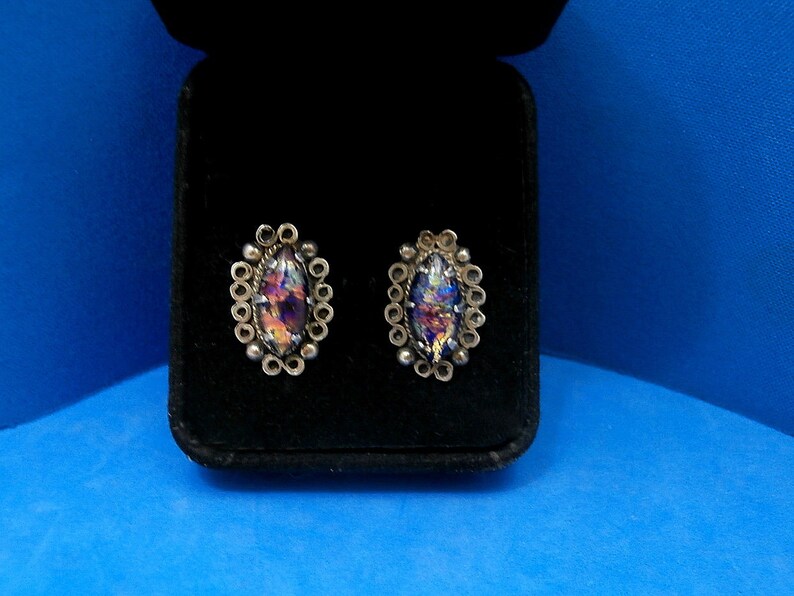 May include: A pair of silver earrings with iridescent glass stones set in a circular design. The earrings are displayed in a black velvet box.