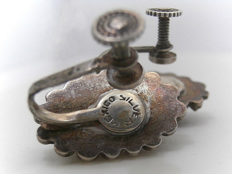 May include: Silver tone earrings with a floral design and a screw back closure. The earrings are marked "T.I.G.O. SILVER".