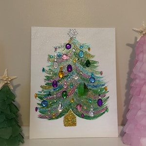 May include: A rectangular canvas featuring a painted Christmas tree decorated with multi-colored glitter and various faceted plastic gems in shades of purple, green, blue, and pink. A silver star sits at the top, and a gold glitter trunk is at the base.