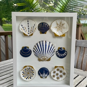 May include: A white framed art piece featuring eight painted seashells. The shells are white, navy blue, and gold, with various dot and line patterns. The frame is white, and the background is a wooden table and greenery.