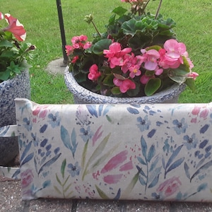 May include: A rectangular fabric bag with a handle, featuring a watercolour floral design in blue, pink, and green hues. The bag is set against a backdrop of potted flowers and greenery.