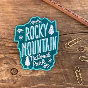 Pode incluir: Um remendo bordado azul turquesa com letras brancas que dizem "Rocky Mountain National Park". O remendo apresenta uma cordilheira de montanhas estilizada e um pinheiro.