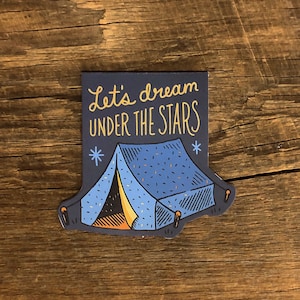 May include: A blue and yellow camping tent sticker with the text "Let's dream under the stars" on a wooden background.