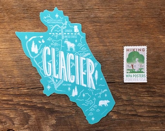 Peut inclure: Un autocollant bleu turquoise du parc national de Glacier dans le Montana avec le texte "GLACIER" en blanc. L'autocollant est sur une surface en bois avec un crayon et un timbre vintage "HIKING".