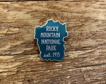 May include: A dark green enamel pin shaped like the outline of Rocky Mountain National Park. The pin has gold lettering that reads "Rocky Mountain National Park est. 1915".