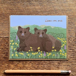 Op de afbeelding: Een wenskaart met twee bruine beren in een veld met gele en paarse bloemen, met de tekst "Lean on me". Een postzegel met gele bloemen staat naast de kaart. Een potlood ligt eronder. Op een houten oppervlak.