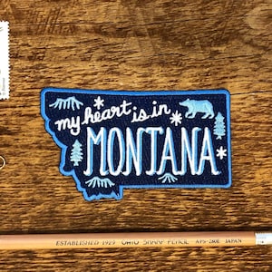 Pode incluir: Patch bordado azul e branco com o texto "my heart is in MONTANA" e uma silhueta de um urso e um pinheiro. O patch tem a forma do estado de Montana.