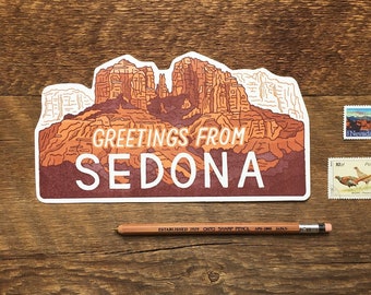 Op de afbeelding: Een witte sticker met een rode en oranje illustratie van een bergketen. De sticker zegt "Greetings from Sedona" in witte tekst.