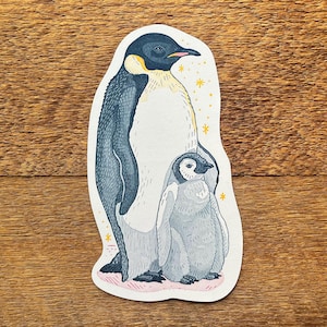 May include: A sticker featuring an illustration of an adult penguin and a chick. The adult penguin is mostly blue and white with a yellow accent. The chick is gray and white. Gold stars are scattered around the penguins.