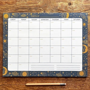 May include: A blue and white monthly calendar notepad with a celestial theme. The notepad has a black border with gold stars, moons, planets, and rockets. The days of the week are labeled: Sunday, Monday, Tuesday, Wednesday, Thursday, Friday, and Saturday. The text "MONTH OF" is at the top of the notepad.