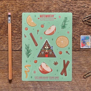Op de afbeelding: Een lichtgroene sticker vellen met vakantie-thema illustraties, waaronder een hut, fruitschijfjes en kruiden. Het vel is gelabeld "NOTWORTHY PAPER & PRESS" en "COZY CABIN HOLIDAY STICKER SHEET". Een potlood, paperclips en een postzegel zijn ook aanwezig.