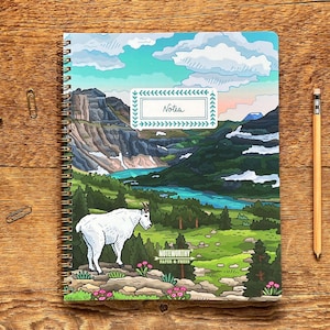 May include: Spiral-bound notebook with a colorful illustration of a mountain landscape, a white goat, and the word "Notes". A pencil rests beside the notebook. The notebook is on a wooden surface.