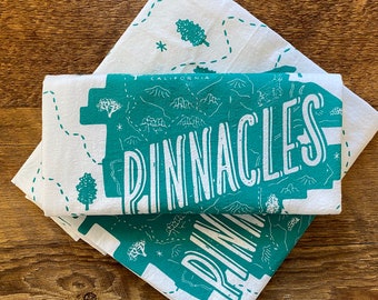 May include: Two white tea towels with a teal green design of a map of the Pinnacles National Park in California. The text "Pinnacles" is printed in white on the map.