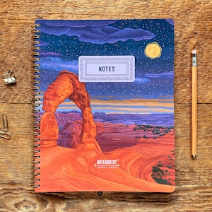 May include: Spiral-bound notebook with a landscape illustration of a desert arch under a starry night sky. The cover features the word "NOTES" and the brand "NOTWORTHY PAPER & PRESS". A pencil and paper clips are on a wooden surface.