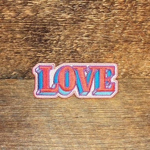 May include: An embroidered patch with the word "LOVE" in a retro style font, featuring red, blue and white colours.