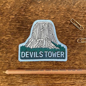 May include: A blue and green embroidered patch with a white outline depicting Devils Tower, a rock formation in Wyoming, USA. The text "Devils Tower" is embroidered in white on the patch.