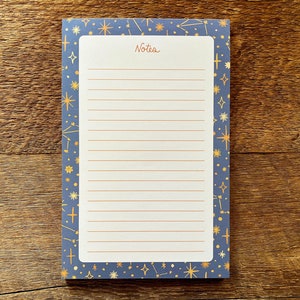 Puede incluir: Un bloc de notas azul con un borde de patrón de estrellas doradas. El bloc de notas tiene papel rayado y la palabra "Notes" impresa en la parte superior.