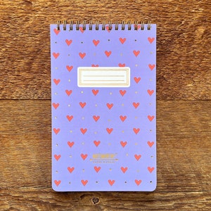 May include: Purple spiral bound notebook with a repeating pattern of red hearts and gold dots. The notebook has a gold label with the text "WRITE NOW" and "PAPER & PEN" on it.