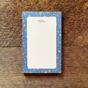 Puede incluir: Un bloc de notas azul con un interior blanco rayado y un borde de estrellas doradas y constelaciones. El bloc de notas tiene la palabra "Notes" impresa en rojo en la parte superior.