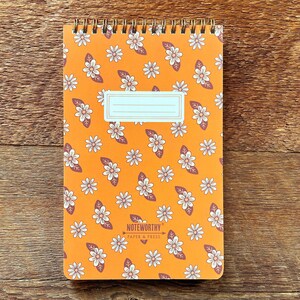 Op de afbeelding: Oranje spiraalnotitieblok met een wit madeliefjespatroon en de tekst "Noteworthy Paper & Press" op de omslag.