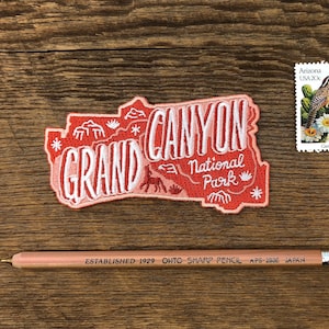 May include: A red and white embroidered patch with the text "GRAND CANYON National Park" and a stylised image of a canyon. The patch is on a wooden surface with a vintage postage stamp featuring a bird and cactus flowers.