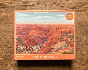 May include: A 1,000 piece jigsaw puzzle featuring a colorful illustration of the Grand Canyon National Park with a mountain lion in the foreground. The puzzle box is orange and features the text "Grand Canyon National Park" and "Noteworthy" with a logo.