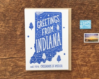 May include: A white card with a blue outline of the state of Indiana. The card says "Greetings from Indiana" and "est. 1816 Crossroads of America".
