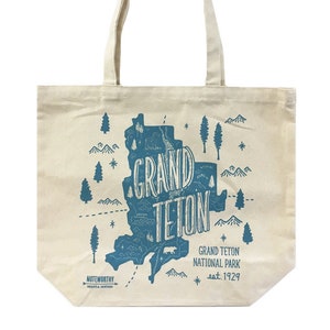 K&ouml;nnte beinhalten: Eine wei&szlig;e Canvas-Tragetasche mit einer blauen Grafik des Grand Teton National Park, Wyoming. Die Grafik enth&auml;lt den Text "Grand Teton" und das Gr&uuml;ndungsdatum des Parks, "est. 1929".