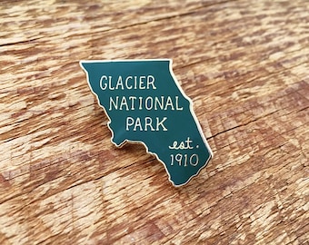 May include: A teal-colored Glacier National Park enamel pin in the shape of the park's outline. The pin features white text that reads "Glacier National Park" and "est. 1910". The pin is set against a wooden background.
