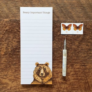 May include: A white notepad with the text "Beary Important Things" at the top. The notepad has lined paper and a brown bear illustration at the bottom. A pen and two butterfly stamps are on a wooden surface.