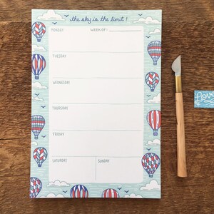 May include: A blue and white weekly planner notepad with a hot air balloon theme. The notepad has the text "the sky is the limit!" at the top and has spaces for each day of the week.