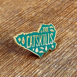 May include: A teal and gold enamel pin in the shape of the Catskills region, with the text "The Catskills" and tree illustrations. Two gold paper clips are on a wooden surface.