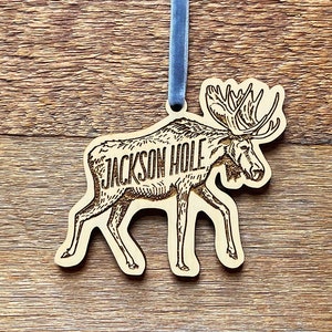 May include: Wooden ornament featuring a moose with the text "JACKSON HOLE" engraved on its side. The ornament is attached to a blue ribbon.