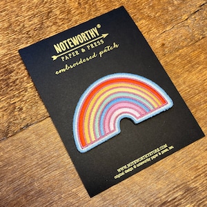 Pu&ograve; includere: Un cerotto ricamato a forma di arcobaleno su una carta nera con il testo "NOTEWORTHY PAPER & PRESS embroidered patch" e "WWW.NOTEWORTHYSTORE.COM original design &copy; noteworthy paper & press, llc".