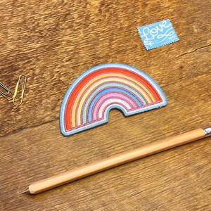 Pu&ograve; includere: Una toppa ricamata a forma di arcobaleno con un contorno blu. La toppa &egrave; su una superficie di legno con una matita e delle graffette.