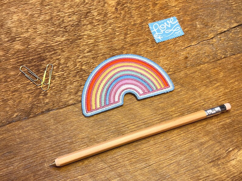 Pu&ograve; includere: Una toppa ricamata a forma di arcobaleno con un contorno blu. La toppa &egrave; su una superficie di legno con una matita e delle graffette.