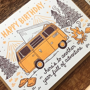 May include: A yellow vintage camper van with a pop-up top drives through a forest with mountains in the background. The card reads "Happy Birthday... here's to another year full of adventure".