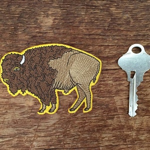 K&ouml;nnte beinhalten: Ein gestickter Aufn&auml;her mit einem braunen und braunen Bison mit gelbem Umriss. Der Aufn&auml;her befindet sich auf einer Holzoberfl&auml;che.