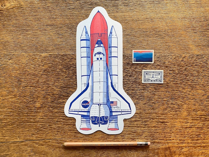 May include: A white and blue space shuttle with red accents, labeled "NASA" and "Atlantis", on a wood background. Two postage stamps are visible in the background.