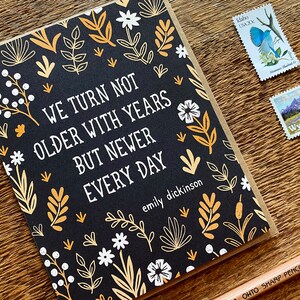 May include: A black card with a gold and white floral pattern. The card has a quote from Emily Dickinson that reads "We turn not older with years but newer every day".