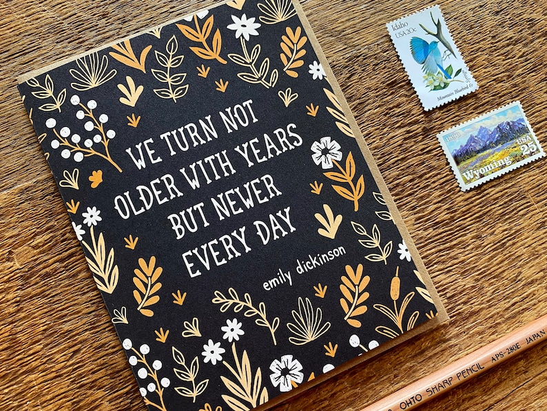 May include: A black card with a gold and white floral pattern. The card has a quote from Emily Dickinson that reads "We turn not older with years but newer every day".