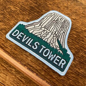 May include: A blue and green embroidered patch featuring a white image of Devils Tower National Monument and the text "Devils Tower" in white.
