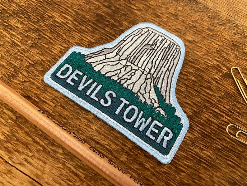 May include: A blue and green embroidered patch featuring a white image of Devils Tower National Monument and the text "Devils Tower" in white.