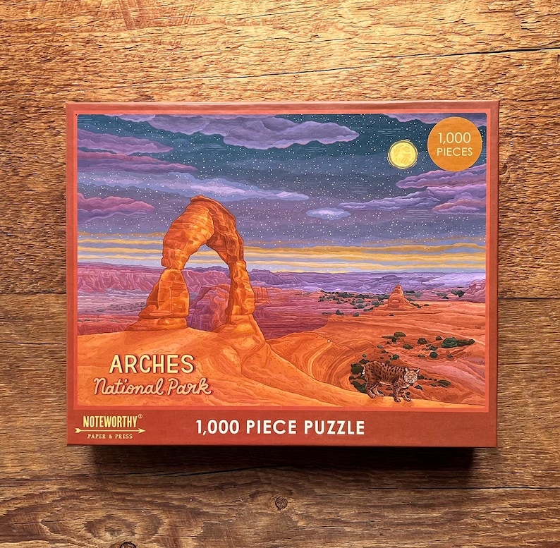 May include: A 1,000 piece jigsaw puzzle featuring a nighttime view of Delicate Arch in Arches National Park, Utah. The puzzle box shows a starry sky with a crescent moon and a lone bobcat in the foreground.