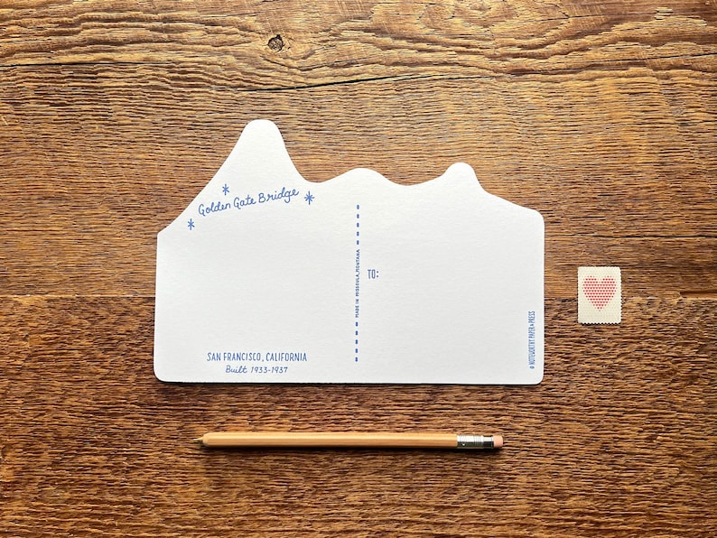 May include: A white postcard shaped like the Golden Gate Bridge with the text "Golden Gate Bridge" and "San Francisco, California Built 1933-1937". The postcard has a blank space for writing a message and a space for the recipient's address.