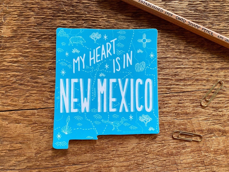 May include: A blue sticker shaped like the state of New Mexico with a white outline. The sticker has a white design of a map of New Mexico with the text "My heart is in New Mexico".
