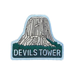 May include: An embroidered patch featuring a white outline of Devils Tower National Monument in Wyoming, with the text "DEVILS TOWER" in white on a green background.