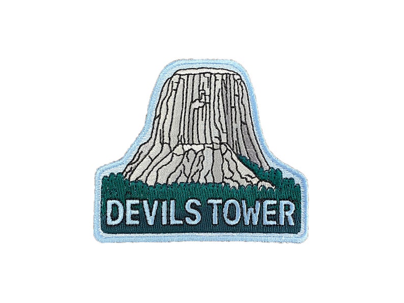 May include: An embroidered patch featuring a white outline of Devils Tower National Monument in Wyoming, with the text "DEVILS TOWER" in white on a green background.