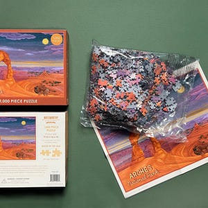 May include: A 1,000 piece jigsaw puzzle featuring a scenic view of Delicate Arch in Arches National Park, Utah. The puzzle box shows a colorful sunset sky with a red rock archway. The puzzle is made in the USA.